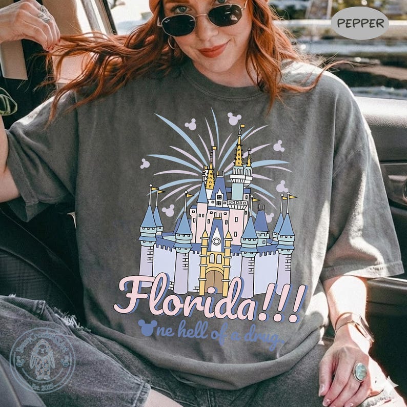 Puede incluir: Camiseta gris con una colorida ilustraci&oacute;n de un castillo, fuegos artificiales y la palabra "Florida!!!" en una fuente l&uacute;dica. El dise&ntilde;o incluye iconos de Mickey Mouse, creando un tema caprichoso.