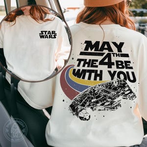Puede incluir: Camiseta crema de Star Wars con el logo de Star Wars en la parte delantera y un gráfico en la parte trasera. El gráfico trasero dice "May the 4th Be With You" con una ilustración de nave espacial y un diseño de arcoíris.