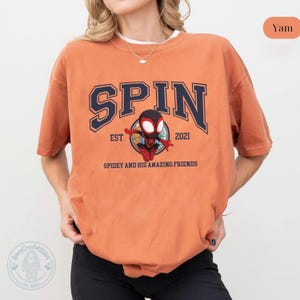 May include: A yam-colored t-shirt featuring the word "SPIN" in navy blue collegiate lettering, with "EST 2021" above a Spidey graphic. The graphic reads "SPIDEY AND HIS AMAZING FRIENDS".