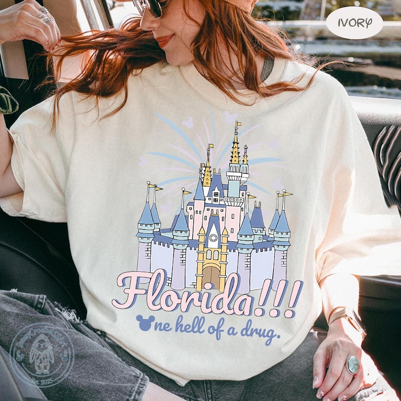 Puede incluir: Camiseta de color marfil con un dise&ntilde;o de castillo de dibujos animados y el texto "Florida!!!" y "one hell of a drug." El castillo es azul, rosa y amarillo. La camiseta tiene un corte holgado.