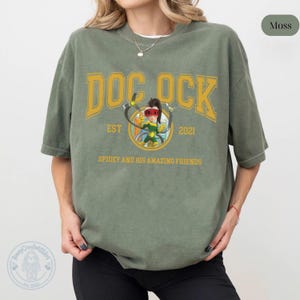 May include: Moss green t-shirt featuring a graphic of Doc Ock from Spidey and His Amazing Friends. The design includes the text "DOC OCK" in yellow, with "EST 2021" and the show's title below. The shirt is a relaxed fit.