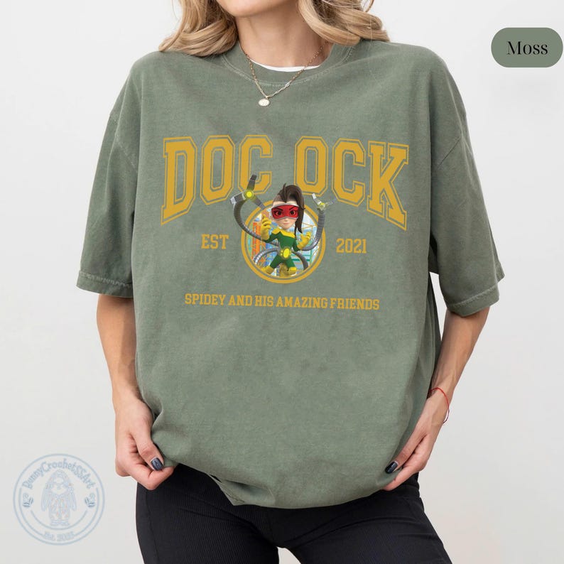 May include: Moss green t-shirt featuring a graphic of Doc Ock from Spidey and His Amazing Friends. The design includes the text "DOC OCK" in yellow, with "EST 2021" and the show's title below. The shirt is a relaxed fit.