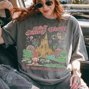 Puede incluir: Camiseta gris con un gr&aacute;fico colorido del castillo de Cenicienta, Mickey Mouse y el texto "Walt Disney World" y "The happiest place in earth". El dise&ntilde;o incluye globos, fuegos artificiales y un autob&uacute;s. La camiseta la lleva una persona.