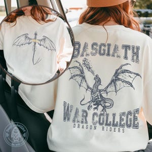 May include: Cream-coloured long-sleeve shirt with a dragon graphic on the front and back. The back design includes the text "BASGIATH WAR COLLEGE DRAGON RIDERS". The dragon illustrations are in a dark grey colour.