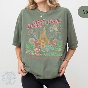 Puede incluir: Camiseta verde musgo con un dise&ntilde;o vintage de Walt Disney World. El dise&ntilde;o incluye un castillo, Mickey Mouse y el texto "The Happiest Place on Earth". La camiseta tiene un corte holgado.