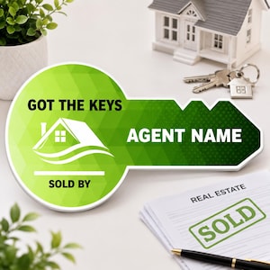 May include: A key-shaped sign with a green geometric pattern, featuring the text GOT THE KEYS, AGENT NAME, and SOLD BY, along with a white house icon. The sign is displayed alongside a miniature house model, metal keys, and a document with SOLD text.