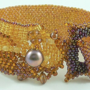 Op de afbeelding: Een kralenarmband met een geometrisch ontwerp in tinten bruin, goud en paars. De armband heeft een grote parel en een vierkant ontwerp.