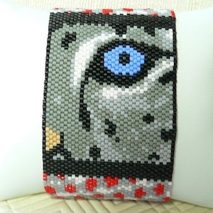 May include: A beaded bracelet with a pixelated image of a blue eye. The bracelet is made with gray, black, white, red, and blue beads.