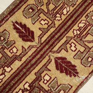 May include: Close-up of a beige and burgundy rug with geometric and floral patterns. The rug features a central beige field with repeating motifs in shades of brown and burgundy, and a burgundy border.