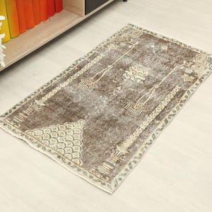 May include: Rectangular brown rug with a distressed, vintage look. The rug features a central design with floral and geometric patterns in beige and light green. The rug is placed on a light-colored wooden floor, with a bookshelf and flowers in the background.