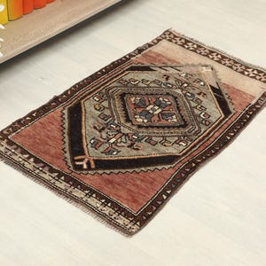 May include: A small, rectangular rug with a central geometric design. The rug features a color palette of muted reds, browns, and grays. The central design is a diamond shape with intricate patterns. The rug has a fringed edge and is suitable for a small space.