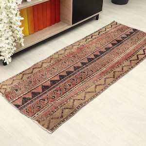 May include: A rectangular, patterned rug with a mix of geometric designs. The rug features alternating stripes of brown, red, and tan, with zigzags and triangle patterns. The rug is placed on a light-colored wooden floor.