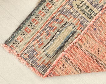 May include: Close-up of a section of a vintage rug with a distressed appearance. The rug features a mix of colors, including coral, blue, beige, and black, with a geometric pattern. The edges are frayed, adding to its antique charm.