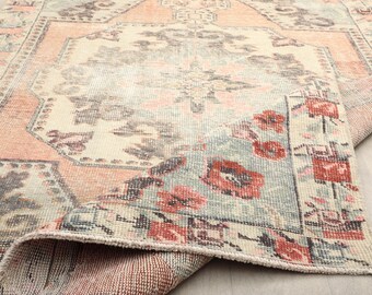 May include: A distressed, vintage-style rug with a rolled edge. The rug features a cream-coloured central field with faded floral patterns in shades of grey and peach. The border and accents showcase red floral motifs and a light blue background.