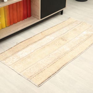 May include: A rectangular beige rug with a striped design. The rug features horizontal stripes in varying shades of beige and tan. It is positioned on a light-coloured wooden floor. A shelf with books and flowers is visible in the background.
