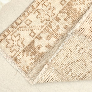 May include: Close-up of a beige and brown patterned rug. The rug has a geometric design in shades of brown and beige. The rug is folded over, revealing the underside with a striped pattern. The edges are frayed.