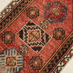 May include: Close-up of a distressed, rectangular rug with a red and black color scheme. The rug features geometric patterns in shades of orange, blue, and brown. The rug's design includes diamond and circular motifs, with a distressed, vintage appearance.