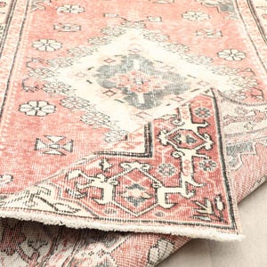 Puede incluir: Una alfombra de estilo vintage y desgastado en tonos coral, crema y gris. La alfombra presenta un patrón central de diamantes y motivos florales, con una sección enrollada que revela la parte inferior y el borde con flecos. El diseño es simétrico.