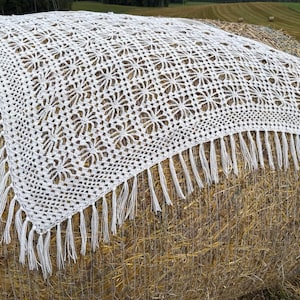 Puede incluir: Un chal blanco tejido a crochet con un patrón floral y flecos. El chal está colocado sobre una paca de heno redonda en un campo. El diseño calado y los flecos añaden textura e interés visual.