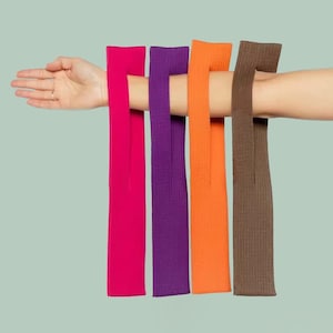 May include: Four colorful arm warmers in fuchsia, purple, orange, and brown are displayed. The ribbed knit arm warmers are shown on an arm against a light green background. The arm warmers are a fashion accessory.