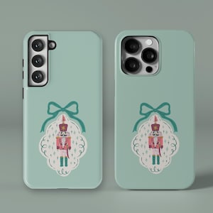 Puede incluir: Dos fundas de teléfono verde menta. Cada funda presenta un diseño decorativo de un soldado cascanueces dentro de un marco blanco y verde azulado, rematado con un lazo verde azulado. El cascanueces es rosa, rojo y dorado.