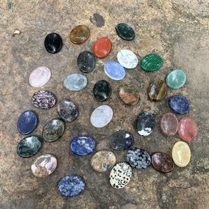 May include: A collection of 25 oval-shaped polished stones in various colors and patterns, including green, blue, red, white, and brown. The stones are arranged on a rough, gray surface.