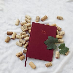 Pode incluir: Um diário bordô intitulado "Vino: A Wine Journal" com uma fita de marcador vermelho escuro, rodeado por rolhas de vinho espalhadas e folhas de uva verdes. As rolhas têm vários desenhos impressos.