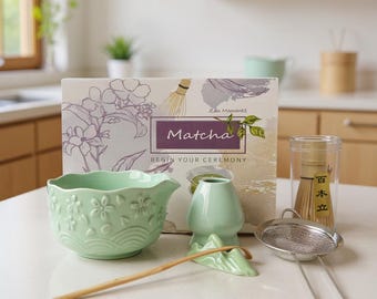 May include: A matcha tea ceremony set featuring a light green ceramic bowl with floral designs, a whisk, a whisk holder, a sifter, and a bamboo scoop. The box in the background reads "Matcha, Begin Your Ceremony."
