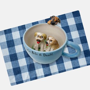 Puede incluir: Una taza de cerámica azul claro con tres figuras de perros. Dos perros están dentro de la taza y uno asoma por el borde. La taza tiene un asa y el texto "Bibi & DanH." escrito en el lateral. Descansa sobre un paño a cuadros azul y blanco.