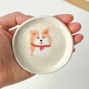 Puede incluir: Un pequeño plato de cerámica blanca con una ilustración pintada a mano de un corgi con un lazo rojo. El corgi está de frente con la lengua fuera.