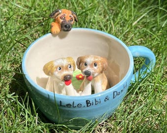 May include: A light blue ceramic mug with three dog figurines. Two dogs sit inside the mug, and one peeks over the rim. The mug is inscribed with "Lele, Bibi & DanH". The background is green grass.