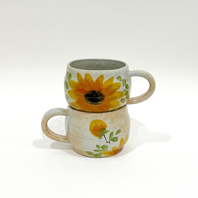 Pu&ograve; includere: Due tazze in ceramica impilate. La tazza superiore presenta un motivo a girasole con petali gialli e un centro marrone scuro. La tazza inferiore ha un motivo floreale giallo. Entrambe le tazze sono di colore beige chiaro.