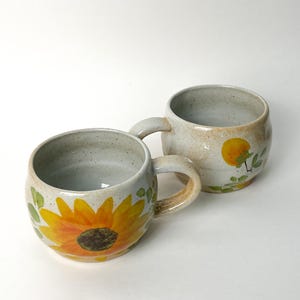 Pu&ograve; includere: Due tazze in ceramica con manici. Una tazza presenta un grande motivo di girasole giallo con un centro marrone e foglie verdi. L'altra tazza ha un motivo di frutta gialla con foglie verdi. Entrambe le tazze hanno uno sfondo bianco sporco maculato.