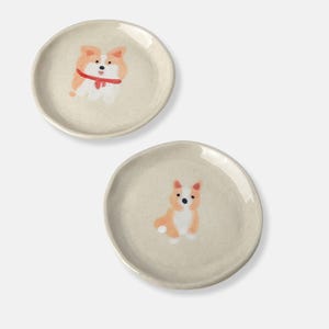 Puede incluir: Dos platos pequeños de cerámica con fondo beige y una imagen pintada a mano de un perro corgi en cada uno.