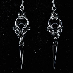 Peut inclure: Une paire de boucles d'oreilles argentées. Chaque boucle d'oreille présente un dessus décoratif avec un motif circulaire, un orbe central et une longue goutte pointue en forme de dague. Les boucles d'oreilles sont suspendues à un simple crochet.