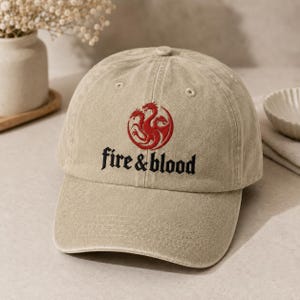 Może przedstawiać: Beżowa czapka z daszkiem o wykończeniu z przecieranego materiału. Z przodu znajduje się wyhaftowany czerwony emblemat trójgłowego smoka nad czarnym napisem "fire & blood" wykonanym gotycką czcionką. Czapka posiada zakrzywiony daszek i otwory.