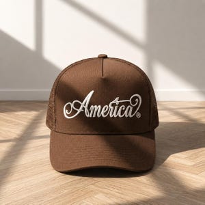 Puede incluir: Gorra de camionero marrón con parte trasera de malla y visera curva. La palabra "America" está bordada en blanco en la parte delantera. La gorra está sobre una superficie de madera.