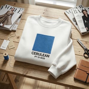 May include: A white sweatshirt with a large, embroidered cerulean blue square and the text "CERULEAN PT 15-4020" below it. The sweatshirt is folded on a wooden surface, with fashion magazines and sewing supplies nearby.