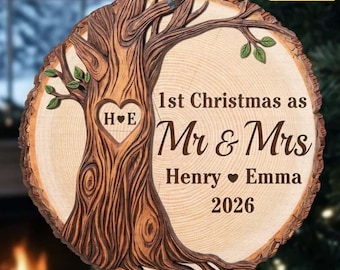 Pode incluir: Um ornamento redondo de madeira com um design de tronco de árvore esculpido e um coração contendo as iniciais H e E. O texto diz 1st Christmas as Mr & Mrs Henry and Emma 2026. Um cordão dourado está preso no topo para pendurar.