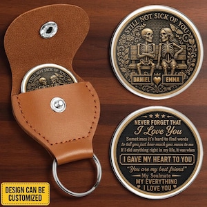 Könnte beinhalten: Ein beigefarbenes Leder-Schlüsselanhänger-Etui mit silbernem Druckknopf und einer runden Metallmünze. Die Münze zeigt zwei Skelette auf einer Bank mit dem Text STILL NOT SICK OF YOU, DANIEL und EMMA. Die Rückseite trägt den Text NEVER FORGET THAT I LOVE YOU.