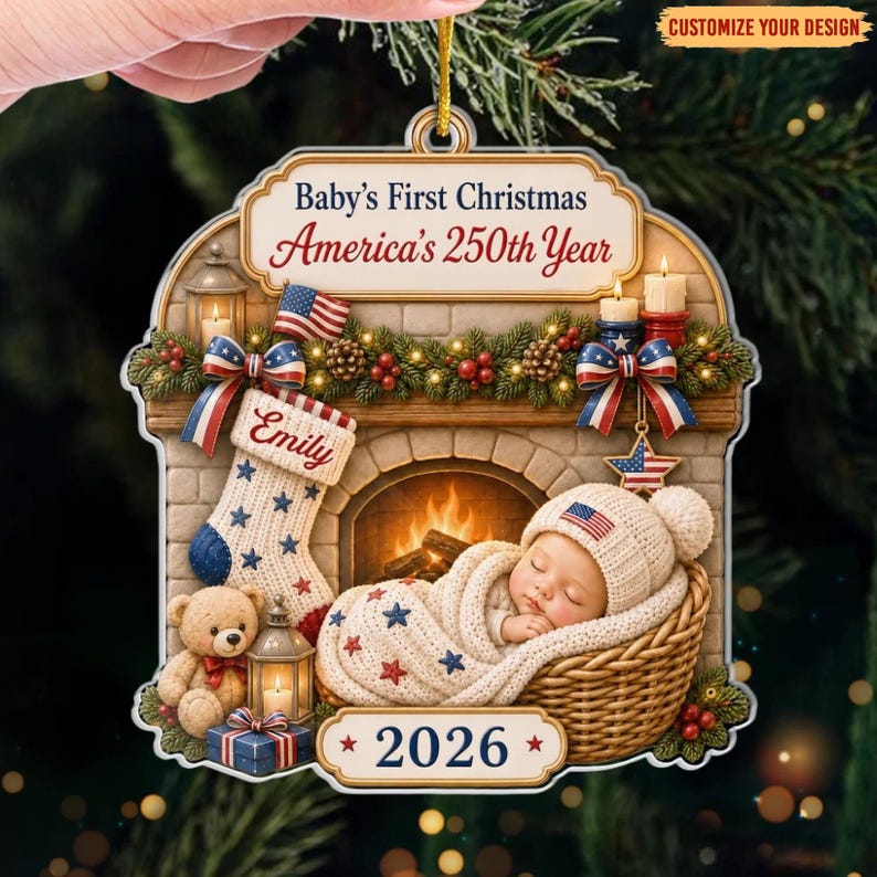 May include: A decorative Christmas ornament featuring a fireplace scene with a sleeping baby in a wicker basket, a teddy bear, and a stocking labeled Emily. The text reads Baby's First Christmas, America's 250th Year, and 2026, with American flag motifs.
