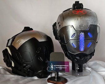 May include: Two futuristic helmets, one black and silver, the other primarily silver with a red star emblem. Both have intricate designs, including blue LED lights and angular shapes. The helmets are displayed on stands, suggesting they are props or collectibles.