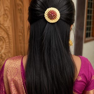 Op de afbeelding: Een cirkelvormig gouden haaraccessoire met een gelaagd, bloembladachtig ontwerp en een cluster van kleine, donkerrode stenen in het midden. Het sieraad zit in lang, donker haar, gecombineerd met een magenta kledingstuk met gouden patronen.