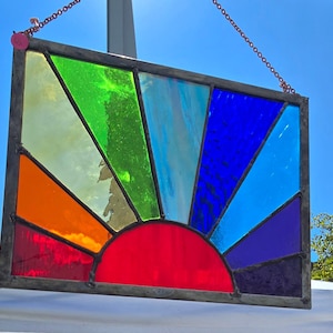 May include: A rectangular stained glass panel with a sunrise design. The sun is a semi-circle of red glass at the bottom, with rays of orange, yellow, green, blue, and purple glass radiating upwards. The panel is framed in a dark metal.