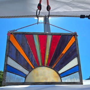 May include: A rectangular stained glass panel showing a sunrise design. It has a semi-circular yellow sun at the bottom, with colourful rays in red, orange, blue, and purple. The panel is framed in dark metal.