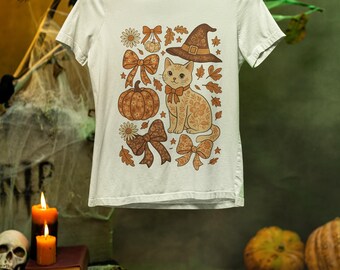 May include: White t-shirt featuring a Halloween-themed design. The graphic includes a cat, witch hat, pumpkin, and bows in shades of orange and brown. The design is surrounded by leaves and flowers.