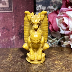 May include: A detailed, yellow-toned sculpture of a dragon-like creature with outstretched wings and a textured body. The figurine has a detailed face and is standing on a round base. The sculpture is a decorative item.