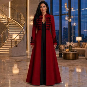 May include: A long, deep red coat featuring black braided military-style frog closures on the front and cuffs, worn over a simple black jumpsuit. The coat has a structured collar and a floor-length hem, creating a bold, uniform-inspired silhouette.