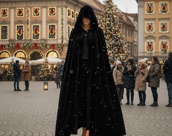 May include: A long, black velvet cloak with a hood, worn outdoors in a snowy environment. The cloak is floor-length and has a flowing design. The background includes a Christmas tree and buildings with festive decorations.