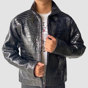 Puede incluir: Una chaqueta de cuero de cocodrilo negro con cierre de cremallera. La chaqueta tiene un cuello de pie y un patrón texturizado.